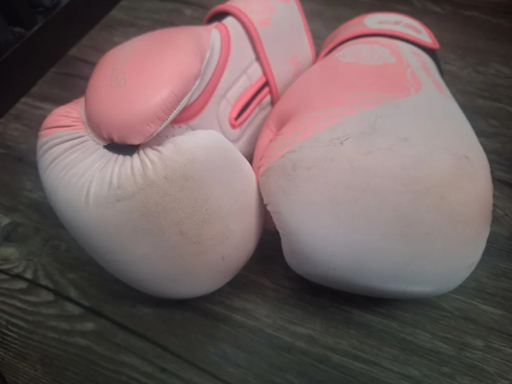 Zwei rosé-weiße Boxhandschuhe nebeneinander auf dunklem Holzboden, Knöchelflächen zur Kamera, Verschmutzung auf beiden Trefferflächen sichtbar